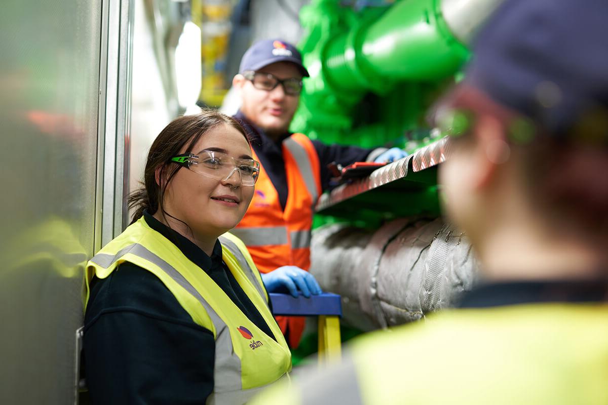 Female engineer in Mitie branded hi vis and safety glasses, standing next to large pipes